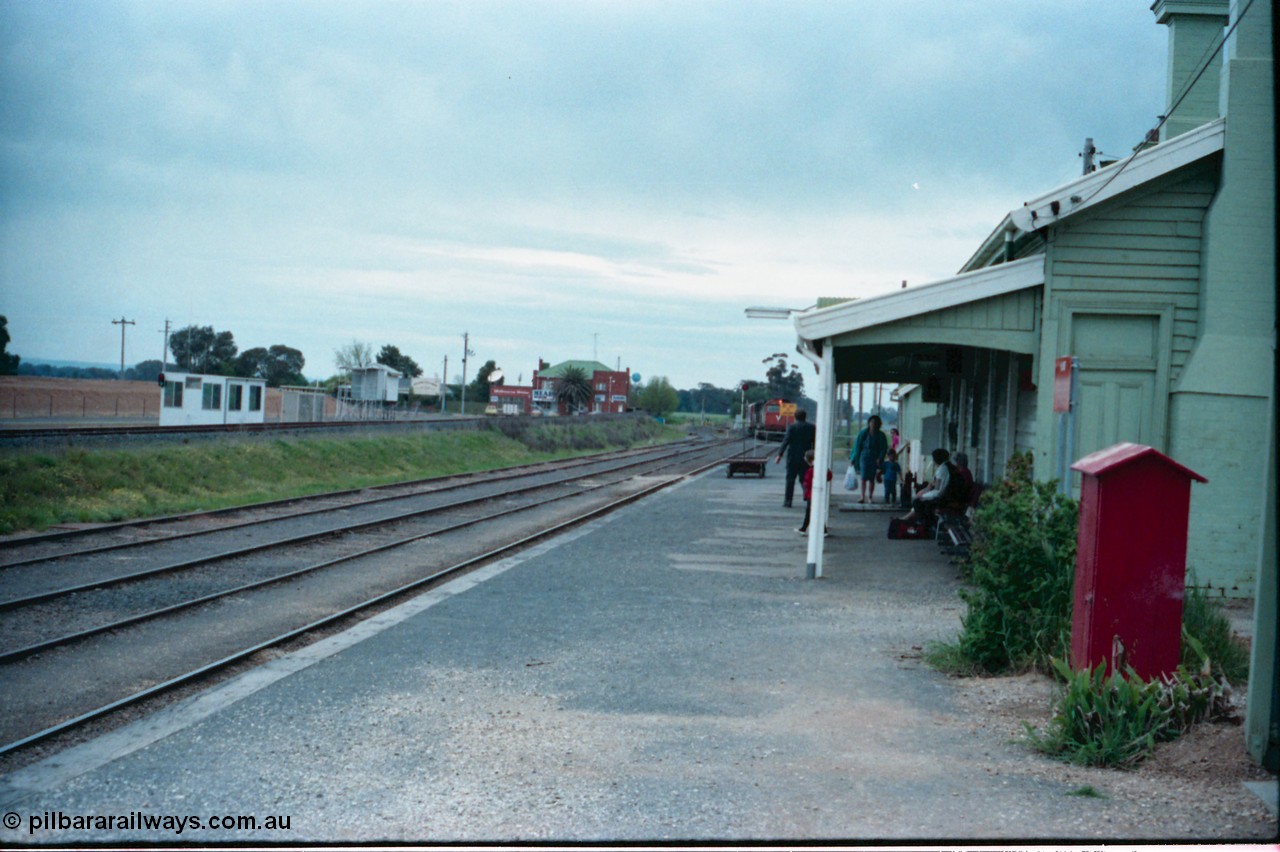 160-07
Murchison East, station platform view of down Cobram passenger train arriving into No.1 Rd, grain receival infrastructure on the left with silos gravitational road, Railway Hotel in the background, passengers awaiting train arrival, station building.
