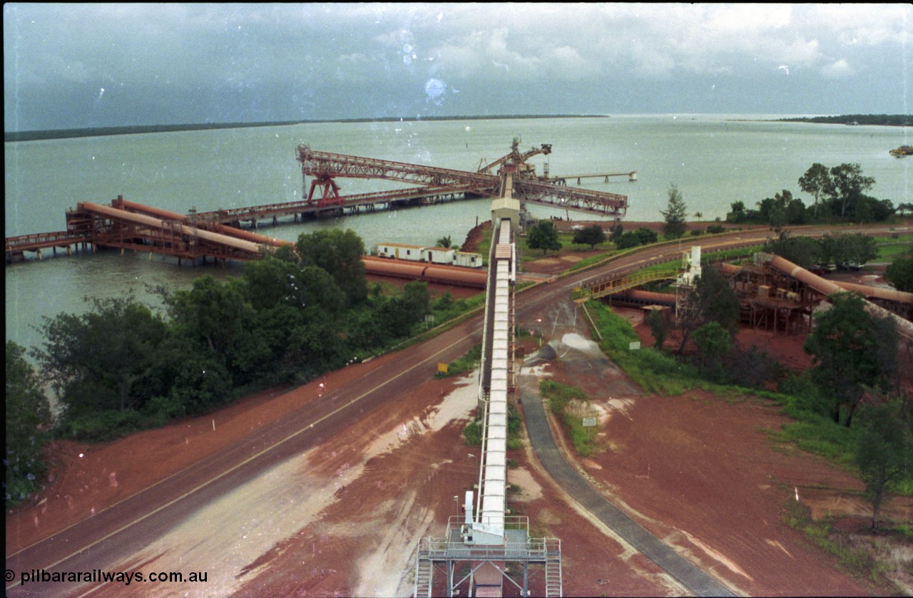 211-00
Weipa, Lorim Point, view from the top of the kaolin storage silos looking north at the kaolin conveyor and ship loader, with red legs and the No.2 bauxite loader straight ahead in the cyclone tie-down position. The bauxite conveyors can be seen passing under the road.
