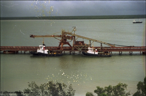 211-01
Weipa, Lorim Point, view from the top of the kaolin storage silos looking west at No.1 bauxite ship loader and the two tug boats. The unloading barge can be seen in the background in the Embley River.
