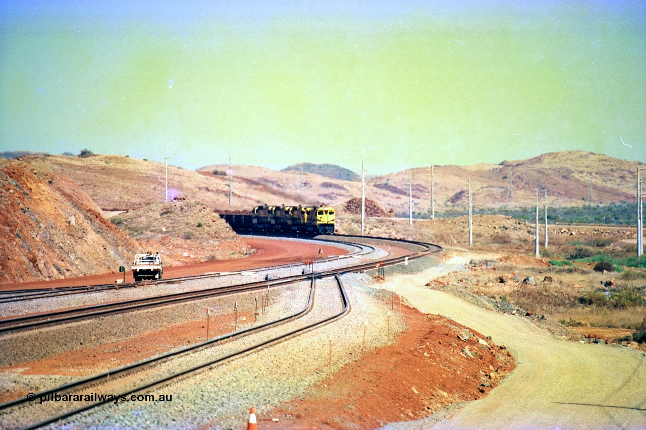 246-07
Cape Lambert, yard view of the then new extension to the south for the West Angelas mine was coming on stream as a Robe River loaded Deepdale train arrives on the main behind the standard quad Dash 8 power with 202 waggons, the just visible siding (rejoining) on the far left will become a compressor waggon holding road, the 6 km and Boat Beach Rd grade crossing are around the corner to the left. Closest to the camera is the extended No. 1 Road which becomes the Empty Car Line from car dumper one for the Deepdale traffic. Next is the original line or Mainline and the third line across is the Loaded Car Line for the West Angelas traffic to the new car dumper 2. 22nd May 2002.
