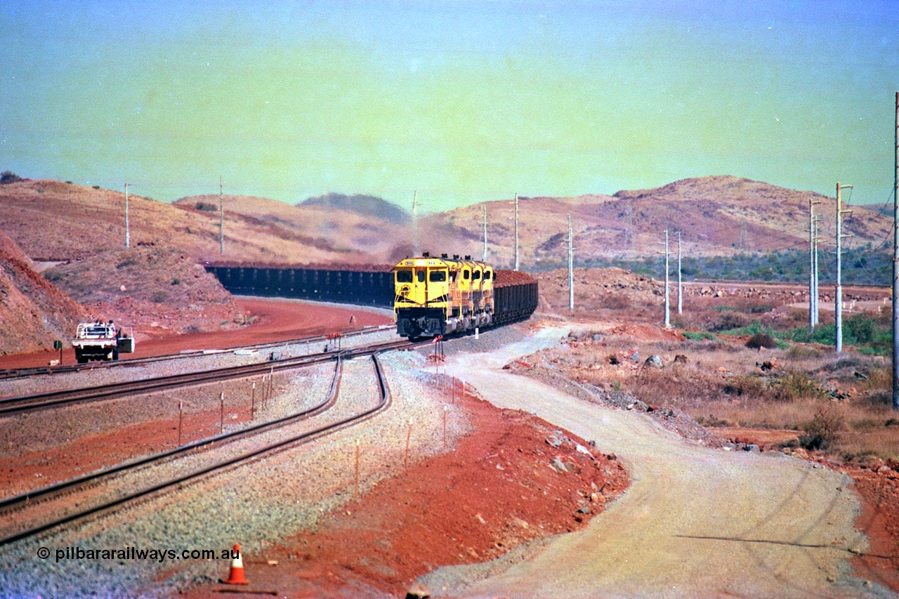 246-11
Cape Lambert, yard view of the then new extension to the south for the West Angelas mine was coming on stream as a Robe River loaded Deepdale train arrives on the main behind the standard quad Dash 8 power with 202 waggons, the just visible siding (rejoining) on the far left will become a compressor waggon holding road, the 6 km and Boat Beach Rd grade crossing are around the corner to the left. Closest to the camera is the extended No. 1 Road which becomes the Empty Car Line from car dumper one for the Deepdale traffic. Next is the original line or Mainline and the third line across is the Loaded Car Line for the West Angelas traffic to the new car dumper 2. 22nd May 2002.
