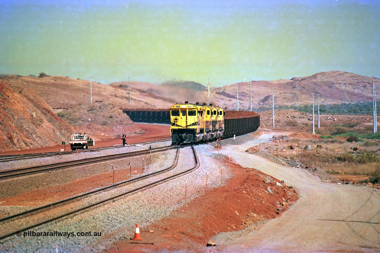 246-12
Cape Lambert, yard view of the then new extension to the south for the West Angelas mine was coming on stream as a Robe River loaded Deepdale train arrives on the main behind the standard quad Dash 8 power with 202 waggons, the just visible siding (rejoining) on the far left will become a compressor waggon holding road, the 6 km and Boat Beach Rd grade crossing are around the corner to the left. Closest to the camera is the extended No. 1 Road which becomes the Empty Car Line from car dumper one for the Deepdale traffic. Next is the original line or Mainline and the third line across is the Loaded Car Line for the West Angelas traffic to the new car dumper 2. 22nd May 2002.
