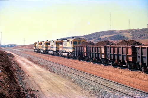 246-27
Cape Lambert, a Robe River loaded Deepdale train rolls along the mainline behind the standard quad Dash 8 power with 202 waggons with newly extended roads either side of it. In the distance can be seen are the railway workshops. 22nd May 2002.
