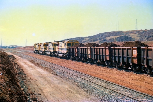 246-28
Cape Lambert, a Robe River loaded Deepdale train rolls along the mainline behind the standard quad Dash 8 power with 202 waggons with newly extended roads either side of it. In the distance can be seen are the railway workshops. 22nd May 2002.
