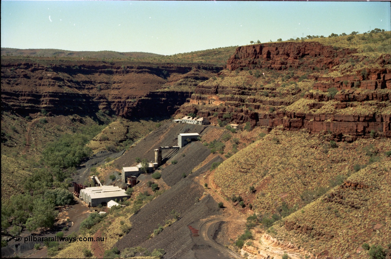 195-16
Wittenoom, view from the Devil's Staircase of Colonial Mill and Mine with piles of tailings, milling site overview, looking south, railway workshops and underground offices are the higher buildings, then the primary crushing shed, holding silo, down to the mill and then the bag house and associated sheds, tailings line the gorge side.
