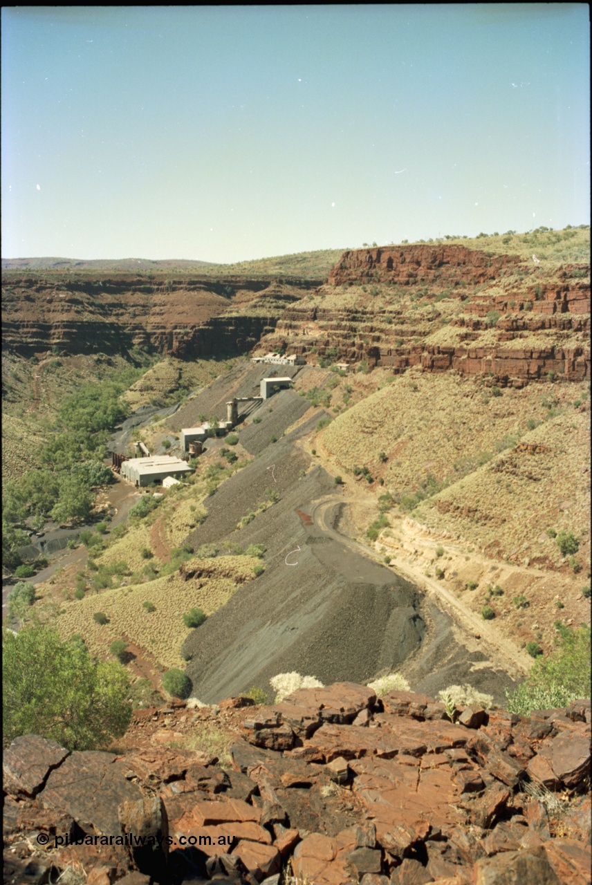 195-22
Wittenoom, view from above the Devil's Staircase of Colonial Mill and Mine with piles of tailings, milling site overview, looking south, railway workshops and underground offices are the higher buildings, then the primary crushing shed, holding silo, down to the mill and then the bag house and associated sheds, tailings line the gorge side.
