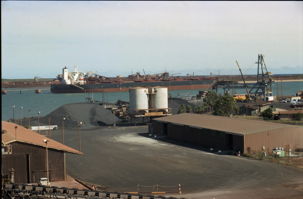 198-28
Port Hedland Port, view of manganese stockpile area with truck unloading, Cockburn Cement silos, old loader can just be made out in front of new bulk loader under construction, 2001.
