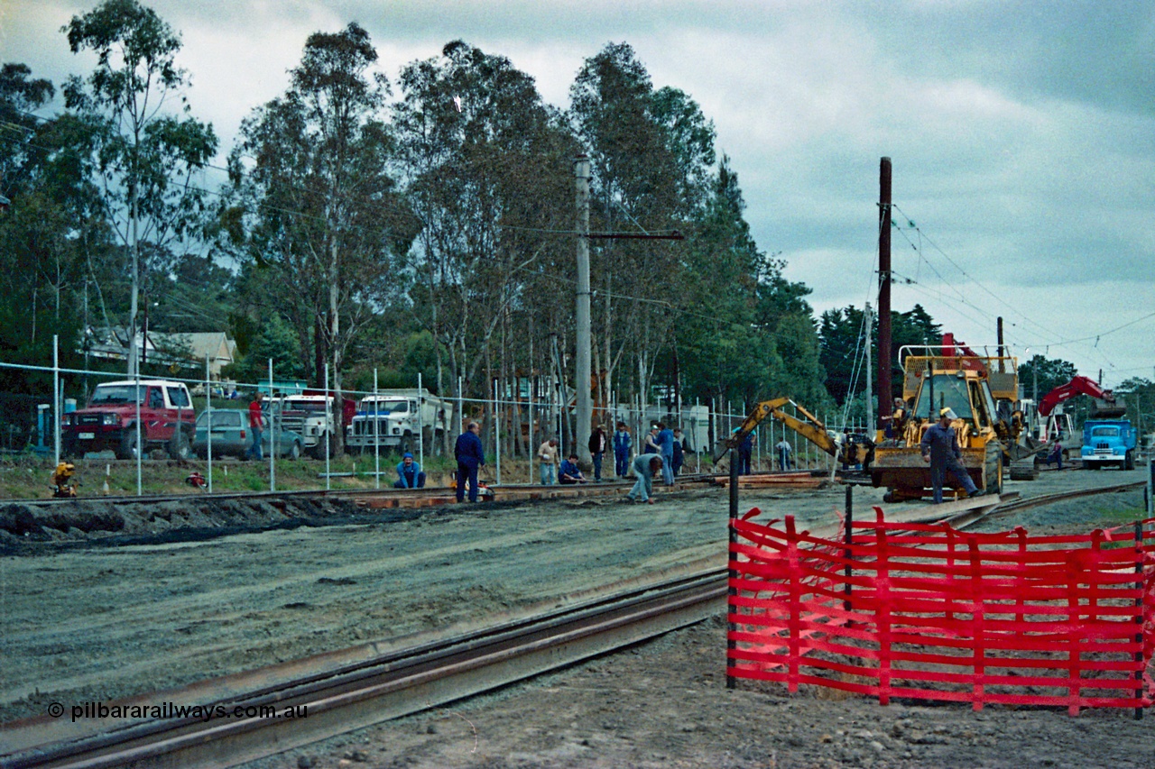 115-01
Hurstbridge, old stabling sidings being removed to make new stabling yard, new fencing, timber traction poles, new sleepers and rail being placed, workers and backhoe, station in the background.
