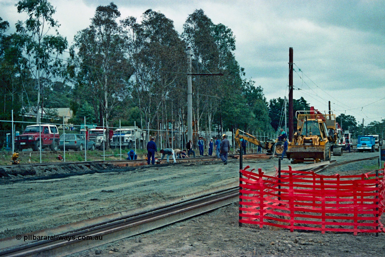 115-02
Hurstbridge, old stabling sidings being removed to make new stabling yard, new fencing, timber traction poles, new sleepers and rail being placed, workers and backhoe, station in the background.
