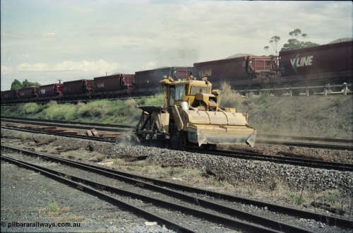 125-10
Seymour, standard gauge mainline, ballast regulator working past loco depot, new and old sleepers along line, broad gauge ballast rake in background.
