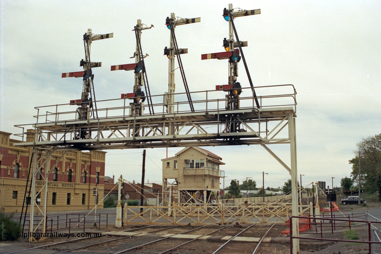 128-04
Ballarat station, view of elevated Ballarat B or Lydiard Street Signal Box, interlocked gates and semaphore signal gantry with semaphore signal Posts 26 and 27 Up signals removed, Post 28 facing away and Post 29, looking west across crib pedestrian crossing, Provincial Hotel on the left.
