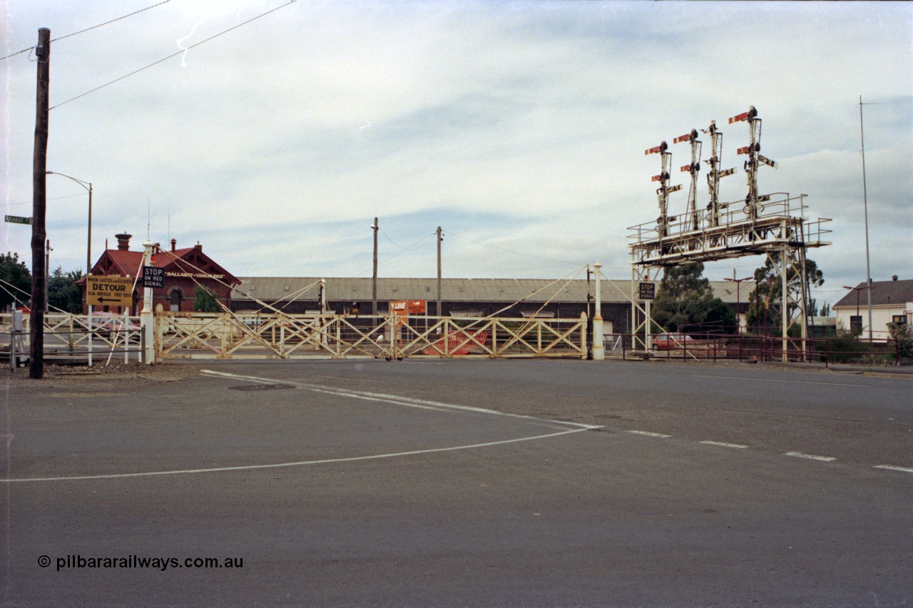 128-14
Ballarat Station, Lydiard St or B signal box, view across Lydiard Street grade crossing with interlocked gates closed, signal gantry with semaphore signal posts 26 to 29, post 26 pulled off for shunt, looking north, Ballarat Coachlines building and Ballarat yard goods shed in the distance.
