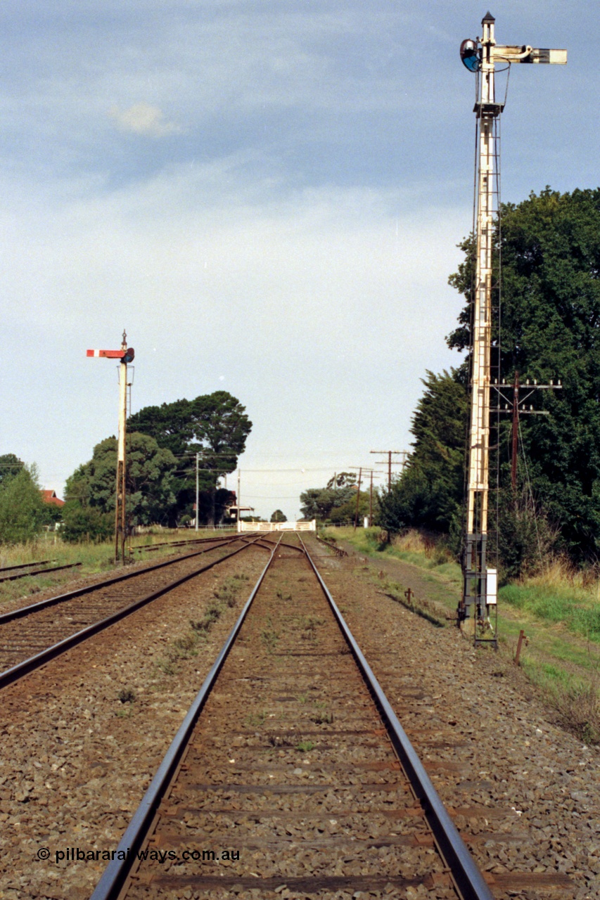 128-18
Gisborne, station overview looking south, semaphore signal Posts 4 and 5, crossover and Up Refuge Siding, Gisborne Road grade crossing non-interlocked swing gates and station in the distance looking towards Melbourne in the up direction.
