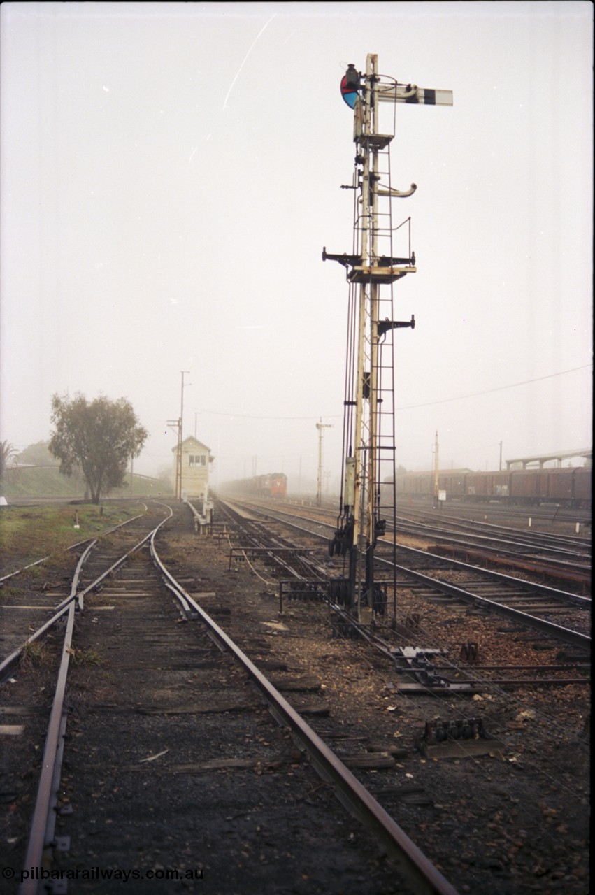 142-1-15
Benalla yard overview, looking beyond a stripped signal post 27, point rodding and wires, tracks to loco depot / workshops at left, Benalla signal box and stabled 9303 goods train in background, very foggy.
