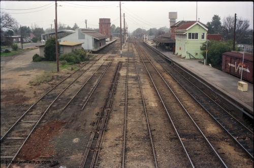 142-2-01
Wangaratta station yard overview looking north from footbridge, No.4 Rd disconnected, goods shed and platform and Freightgate canopy, water tower and crane on the left, station building and elevated signal box on the right, north end footbridge visible in the distance, point rodding running down middle of yard.
