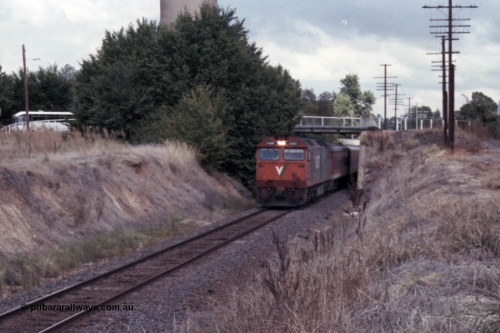 148-30
Wangaratta, V/Line standard gauge G class G 520 Clyde Engineering EMD model JT26C-2SS serial 85-1233 leads the down Inter-Capital Daylight under the station access bridge with town water tower behind bushes, broad gauge yard and station is to the right of image.
