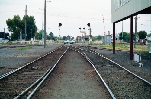 156-07
Shepparton yard view looking north across High Street grade crossing from in-between No.1 and 2 roads, former 'engine track' is on the right, points locked for Tocumwal line on the left, line on the right is to Katamatite, the points on the left of frame are from No.5 Road, searchlight signal posts 10 and 11 are the down home signals for each line, track on the far left cross High Street and is the former SEC Siding, SPC Siding can be seen on the right in the distance. Awning at right is the former J. Murray More Pty Ltd or Tubemakers unloading gantry.
