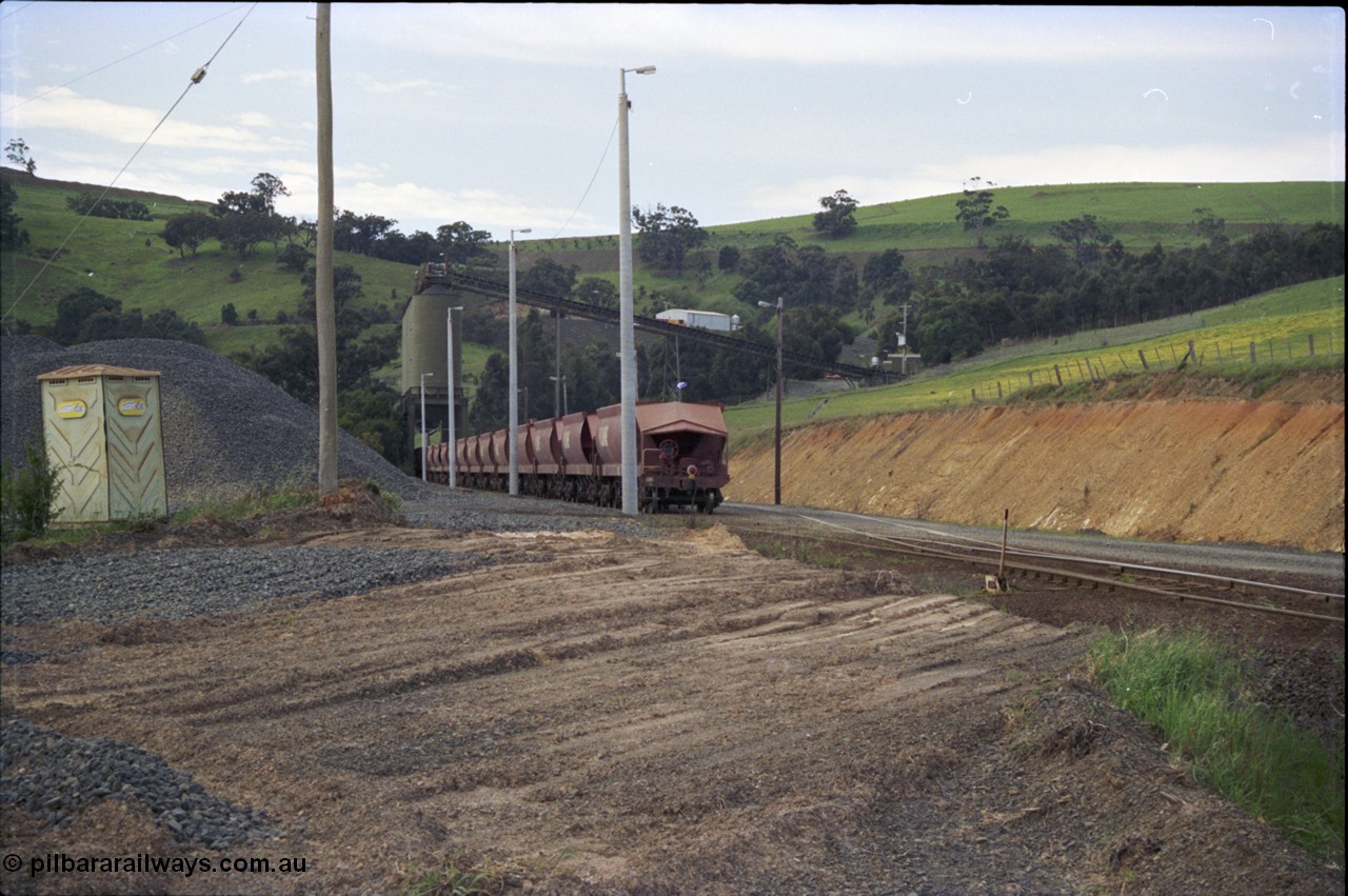 157-07
Kilmore East, Apex Quarry Siding, empty broad gauge V/Line stone train rake is under the loading bins having just arrived, locomotives cutting off to shunt round, oversize ballast pile and 'Super Loo' on the left.
