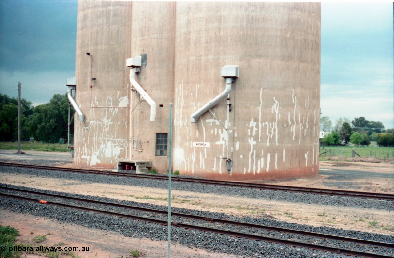 161-11
Katunga, view across tracks from former station site, location sign missing from post, station name painted on Williamstown style silo complex, three train loading spouts.
