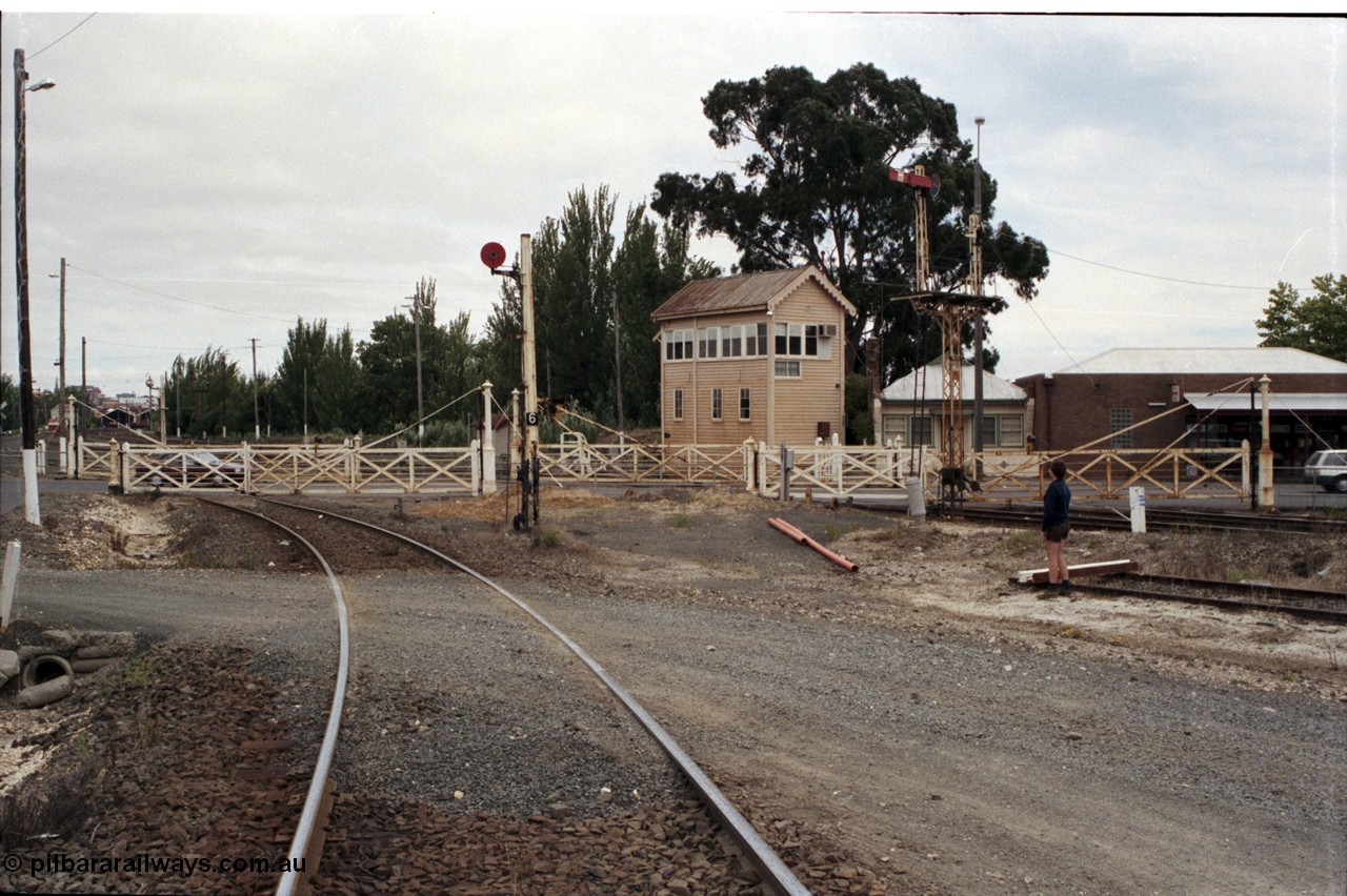 172-36
Ballarat East signal box, track view looking across interlocked swing gates at Humffray St, looking west towards Ballarat, signal box framed between semaphore signal post 5 and disc signal post 6. Disc signal 6 is down home from Loco Tracks to Post 15 controlled by A Box, Post 5 left doll is Down Home No. 2 to Post 11 via Passenger Line controlled by A Box.
